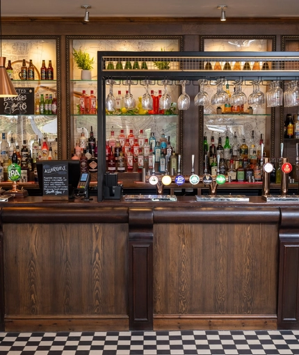 A close up view of the bar at The Man On The Moon, showing the beer taps, bottles of various wines and spirits on shelves, and glasses hanging in racks above the bar.