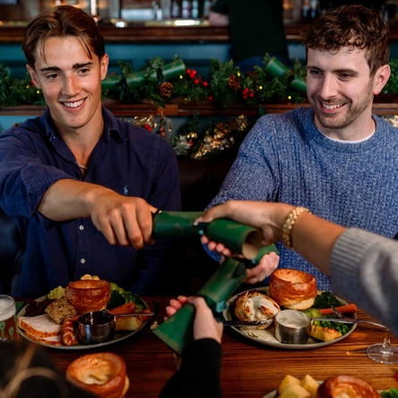 An image focusing 2 people sat within a group at a table within the interior restaurant seating area pulling a cracker while enjoying drinks and dishes from the Christmas menu at a Community Social venue.
