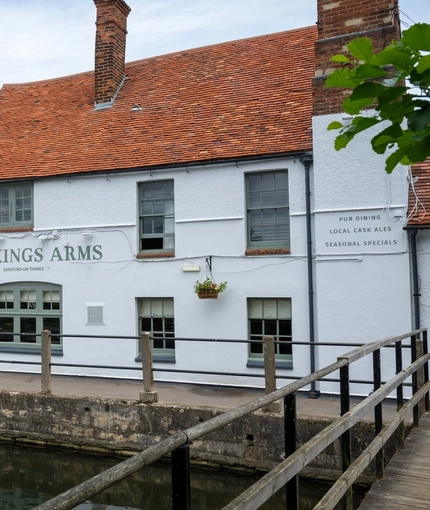 A view from across the river of the exterior facade of The Kings Arms in Sandford-On-Thames, and the wooden bridge leading across the water.