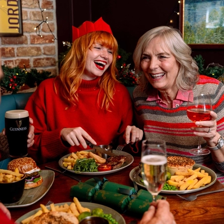 Two people seated together in a festively decorated pub booth, looking towards each other across a table set with seasonal meals. One person wears a red paper crown and red jumper while cutting into a steak meal served with chips, rocket and a grilled tomato. The other holds a glass of rosé beside a pie meal with chips and vegetables. Burgers, drinks and green Christmas crackers are arranged on the table, with Christmas garlands, fairy lights and a brick wall creating a warm festive pub setting.