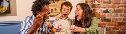 Two adults and a child are seated around a table in a pub, sharing a selection of desserts. The table features a large ice cream sundae, waffles topped with ice cream and sauce, cheesecake with strawberries, a chocolate dessert, a glass of white wine, a blue mug of coffee and a children's juice carton. Exposed brickwork, blue panelled walls and a wooden barrel-style wall sign create a warm and inviting pub setting.