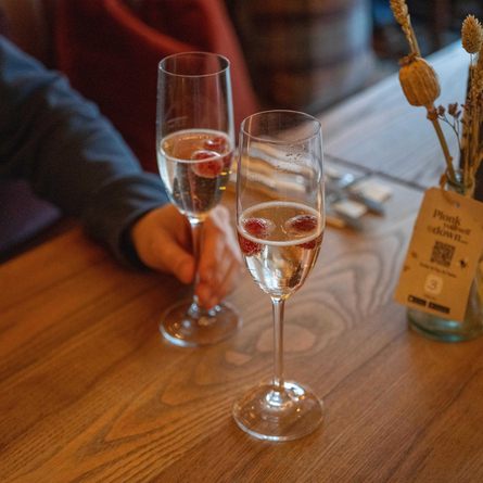 A close up view of two glasses of Prosecco garnished with berries, sitting on a wooden table next to cutlery, a napkin, a menu, and table decorations. A person at the table is holding the stem of one of the glasses.