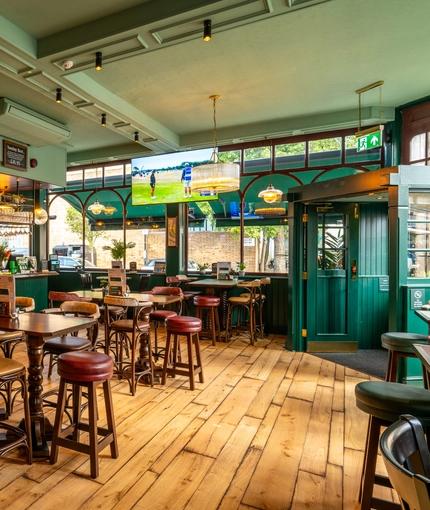 The bar and interior restaurant seating area at the Phoenix, with wooden flooring, bar stool seating, and a large TV hanging on the wall above the windows.