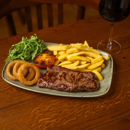 Steak & Chips served on a plate with salad, tomato, and onion rings. The plate sits on a wooden restaurant table along with a glass of red wine.