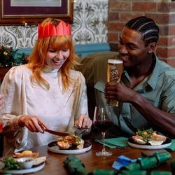 A person wearing a red paper crown sits at a dining table in a festive pub, holding a knife and fork beside a plate of food that includes prawns, potatoes and salad leaves. Christmas crackers, drinks and additional dishes are arranged on the table, while garlands with lights and seasonal decorations line the booth seating in the background.