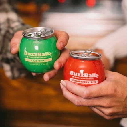 A group of people stood near a bar,  holding cans of  BuzzBallz cocktails.