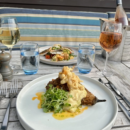A plated aubergine schnitzel and a plated sea bass and tiger prawn salad with wine and water sat on an exterior table at The White Horses.