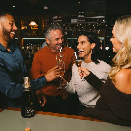 Four people standing at a table in a bar, laughing together and raising glasses of champagne. A bottle of Lanson champagne stands on the table, and a Christmas tree decorated in red and gold stands beside them.