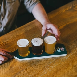 A man is seated at an indoor wooden table which is holding a Beer Flight Paddle with 3 mini pint glasses of beer on it. The man's arms are on the table and he is reaching for one of the glasses.