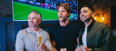A group of friends stood round a table in the seating area in a Greene King pub with drinks watching sports.