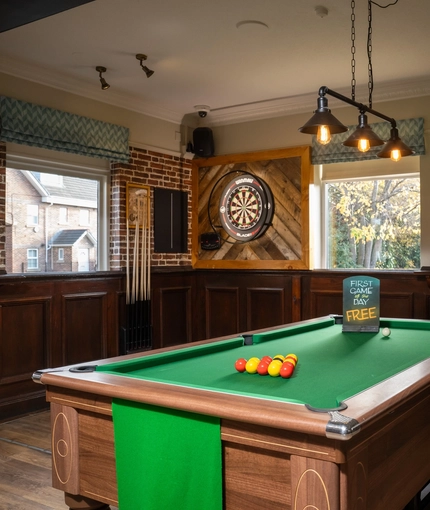 The interior pub gaming area of The Longship, with a dartboard, a pool table, and bar stool seating.