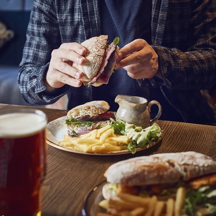 A roast beef sandwich on a table being picked up and eaten, with another sandwich and a pint of Greene King IPA in front of it.