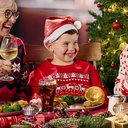 Three adults and a child sitting side by side at a restaurant table and eating from the plates of Christmas dinner in front of them. Glasses of beer and fizzy drink stand on the table along with Christmas presents, Christmas crackers, baubles, and pine cones; two of the adults are holding glasses of wine; and a Christmas tree is visible in the background.