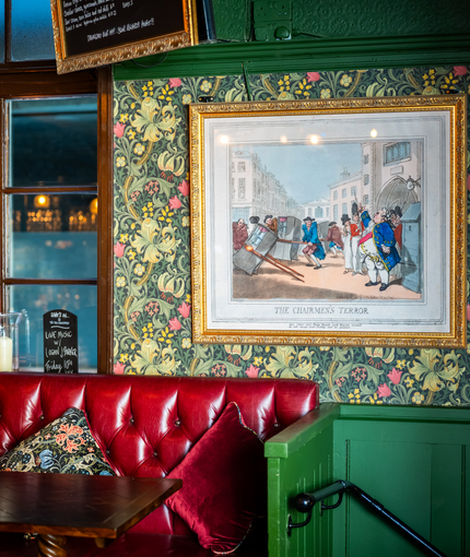 A close up view of a red, upholstered booth seat in front of a window inside the Two Chairmen, with a framed art piece on the floral wallpapered wall next to it.