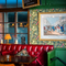 A close up view of a red, upholstered booth seat in front of a window inside the Two Chairmen, with a framed art piece on the floral wallpapered wall next to it.
