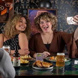 Three people sit at a restaurant table. Two people on the far side of the table sit close together; one holds up a smartphone as if taking a selfie while the other holds a bottled drink and a glass with ice. The table in front of them is filled with food.