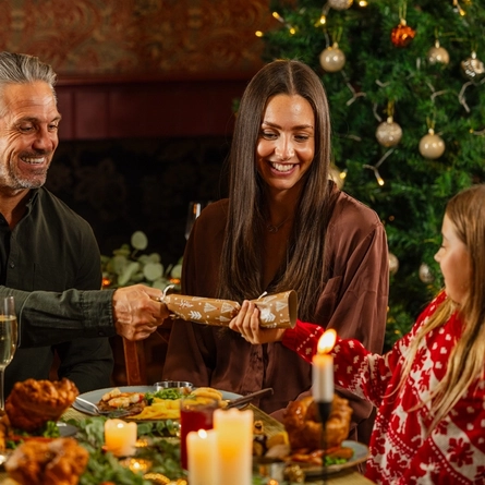 An image of 3 adults and a child pulling Christmas crackers while enjoying main dishes and drinks within the interior restaurant seating area at a Venture Hotel venue.