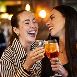 Two women at a table in a pub bar area, laughing together. One of them is holding a glass of champagne, the other is holding a cocktail, and other drinks sit on the table in front of them.