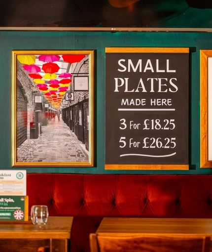A close up view of framed artwork and a chalkboard sign advertising small plates, on a dark green wall above an upholstered booth seat inside the Royal George.