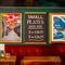A close up view of framed artwork and a chalkboard sign advertising small plates, on a dark green wall above an upholstered booth seat inside the Royal George.