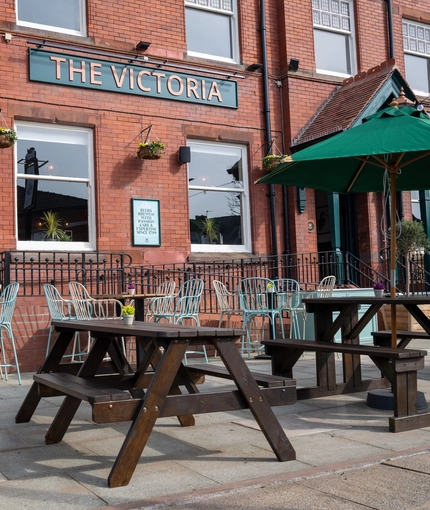 The exterior beer garden seating area at the front of The Victoria, with wooden picnic tables and shade umbrellas.