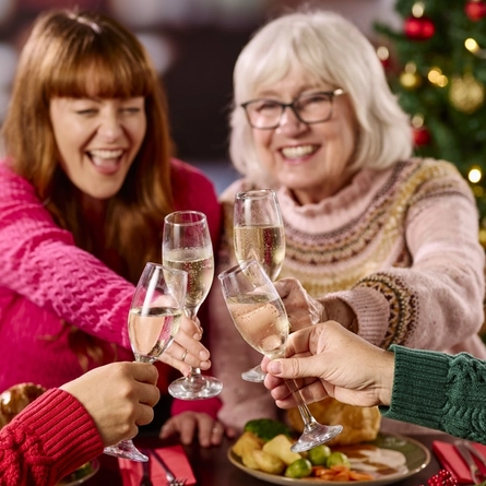 A close up view of four glasses of sparkling wine being clinked together by four people sitting at a restaurant table. Cutlery, serviettes, plates of Christmas dinner, and a tealight sit on the table; and a Christmas tree is visible in the background.