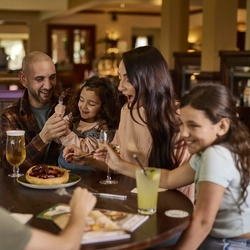Three adults and three children sitting around a wooden restaurant table inside a Farmhouse Inns venue. Glasses of beer, white wine, and a soft drink sit on the table; as well as a plate of pigs in blankets served in a giant Yorkshire pudding.