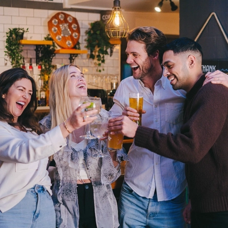 Four people standing beside the bar in a pub with their arms around each other. Two of them are holding glasses of beer, and the other two are holding cocktails.