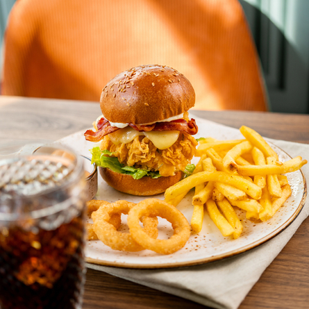 A Crispy Chicken Burger served on a plate with fries, onion rings, and a small pot of dipping sauce. The plate sits on a cloth napkin on a wooden table, along with cutlery, a glass of fizzy drink, and a glass vase filled with dried flowers.