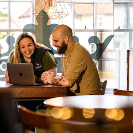 2 staff members sat smiling at a laptop.