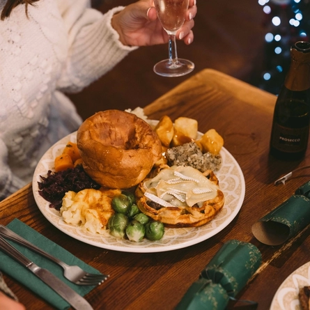Festive vegetarian roast dinner served on a pub table, featuring a pastry tart topped with melted brie, a Yorkshire pudding, roast potatoes, Brussels sprouts, cauliflower cheese, stuffing and red cabbage. A glass of sparkling wine and a miniature bottle of prosecco sit beside the meal, with green Christmas crackers and twinkling Christmas tree lights in the background.