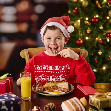 A child sitting at a restaurant table, wearing a Santa hat and eating from the plate of cooked breakfast in front of them. Christmas presents and a glass of juice also sit on the table; and a Christmas tree is visible in the background.