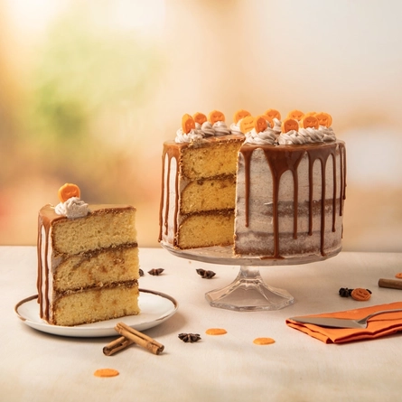 The Pumpkin Spiced Latte cake, served on a glass cake stand which sits on a table strewn with cinnamon sticks, star anise, and mini pumpkin decorations. A slice has been cut out of the cake and served on a plate which sits on the table alongside the cake stand.