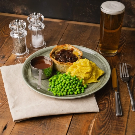 An advertising image showing a plated Croxton Manor Cheddar, Double Gloucester & Leek Pie with mashed potatoes, peas and sauce sat on a wooden table.