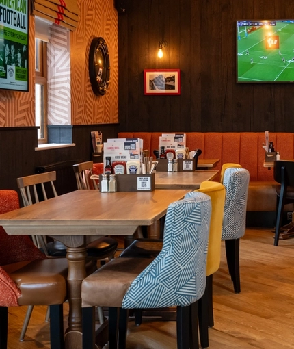 The interior restaurant seating area at the Arch & Anchor, with wooden tables, upholstered seats, and TVs on the walls.