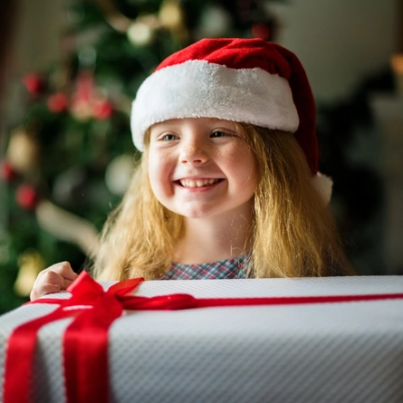 A young girl, wearing a Santa hat, and holding a white present wrapped with a red bow.