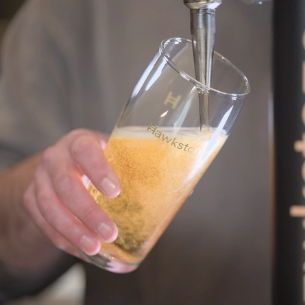 A lifestyle image showing a member of staff stood behind a bar pouring a pint of Hawkstone lager.