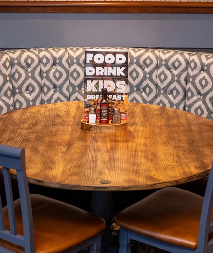 A close up view of an upholstered, curved booth seat and round restaurant table, inside at The Railway. Two more chairs are placed in front of the table, and framed artwork hangs on the wall above the booth seat.