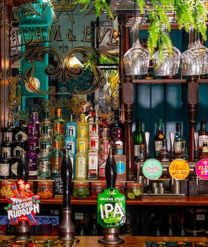 A close up view of the bar inside Adam & Eve in St James, with glasses and artificial plants hanging in a rack above the counter; and bottles of alcohol, jars of nuts, and ornaments sitting on wooden shelves behind the bar.