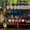 A close up view of the bar inside Adam & Eve in St James, with glasses and artificial plants hanging in a rack above the counter; and bottles of alcohol, jars of nuts, and ornaments sitting on wooden shelves behind the bar.