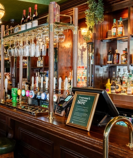 The dark wood panelled bar inside the King's Head in Bayswater, with bar stool seating; dark wood flooring with a checkerboard tile pattern by the bar; and glasses hanging in racks above the counter.