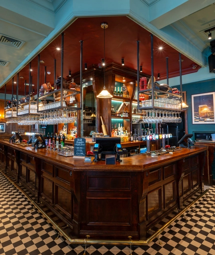 The wood panelled bar, and interior restaurant seating area at the Blue Posts in St James's, with a TV on the wall, and checkerboard flooring around the edge of the bar.