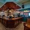 The wood panelled bar, and interior restaurant seating area at the Blue Posts in St James's, with a TV on the wall, and checkerboard flooring around the edge of the bar.
