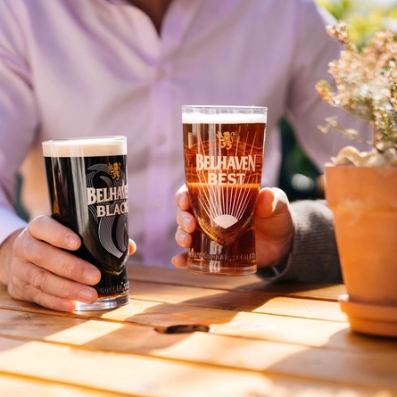 A close up view of two hands holding glasses of Belhaven Black and Belhaven Best, at a wooden table in a sunny beer garden.