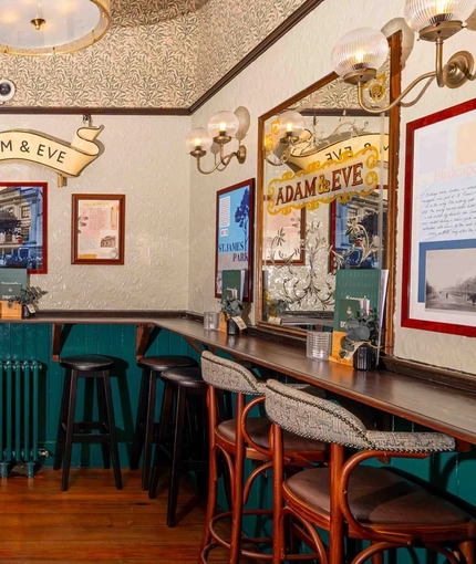 An interior restaurant seating area at Adam & Eve, with bar stool seating, framed artwork on the walls, and potted plants on a shelf.