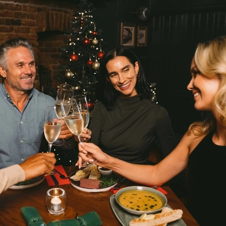 A close up view of four people sitting at a restaurant table laden with plates of food, clinking together glasses of Lanson champagne. Christmas crackers and the bottle of champagne sit on the table, and a Christmas tree decorated in red and gold is visible in the background.