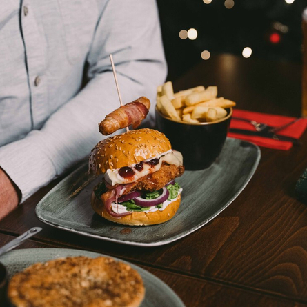 A close up view of a burger served on a plate with a pig in blanket and a bowl of fries. The plate sits on a wooden restaurant table along with a glass of Hazy Day, another plate of food, a Christmas cracker, and cutlery; and a person is sitting at the table.