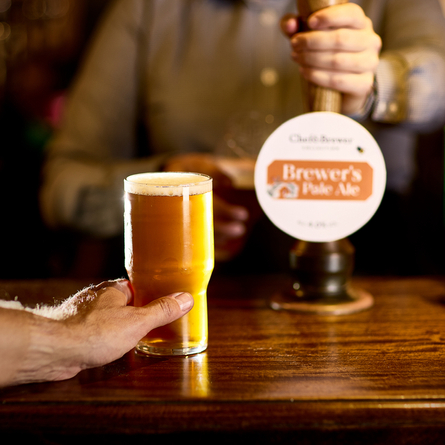 An image of a customer holding a pint of Brewer's Pale Ale stood at the interior bar with a member of staff pulling a pint in the background at a Chef & Brewer venue.