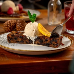 A close-up of a dessert plate featuring a slice of fruitcake topped with a scoop of vanilla ice cream, garnished with a mint leaf and an orange slice. A hand is holding a spoon, ready to take a bite.