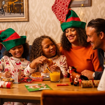 A festive restaurant booth decorated for Christmas, with four people seated around a wooden table. Two people wear green-and-red elf hats, while another wears Christmas-themed pyjamas. Plates of breakfast food, a glass of orange juice, a juice carton, Christmas crackers, colouring materials and a small reindeer toy are arranged on the table. A red Christmas stocking hangs on the patterned wall behind the group, with framed artwork and string lights completing the seasonal setting.